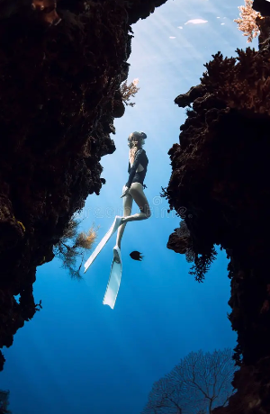 Freediver with long fins descending through clear blue water between coral-covered reef walls.