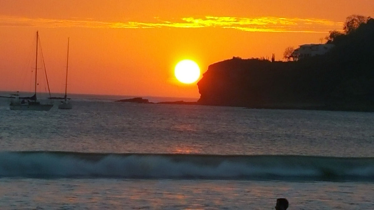 A scenic sunset over the ocean with the sun beside the face of the Indian cliff, sailboats offshore, and a breaking wave in the foreground. 