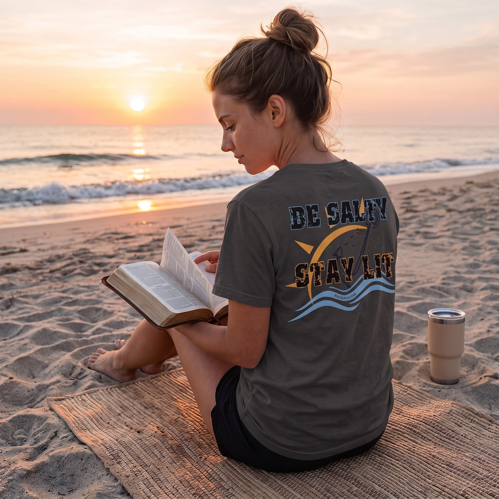 Woman wearing a Dark Grey Deepwater Crew Co. Be Salty, Stay Lit T-shirt, back view while sitting on the beach at sunset.