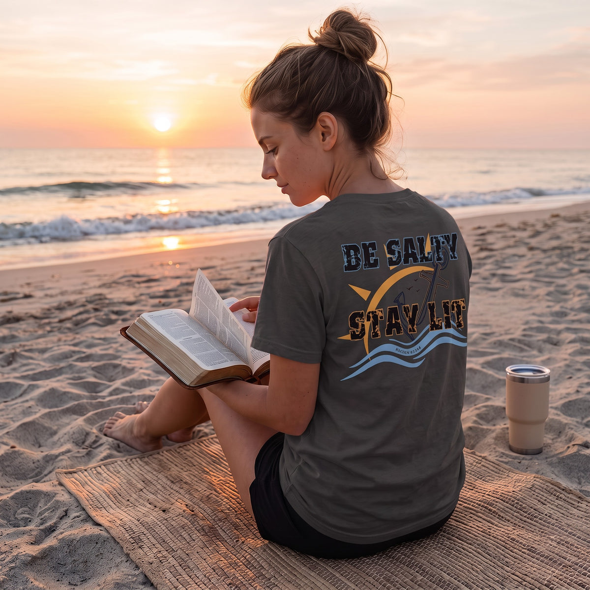 Woman wearing a Dark Grey Deepwater Crew Co. Be Salty, Stay Lit T-shirt, back view while sitting on the beach at sunset.