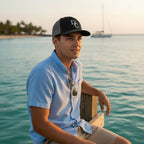 Deepwater Crew Co. black and grey mesh trucker cap with DC logo worn by a man on a wooden pier by the ocean.
