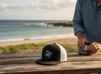 Deepwater Crew Co. black trucker cap with white mesh back and embroidered wave compass logo. Flat bill design.