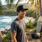 Deepwater Crew Co. black trucker cap with embroidered logo worn by a man on a tropical beach with a surfboard
