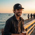 Deepwater Crew Co. black trucker cap with white wave logo worn by a smiling man on a pier at sunset.
