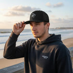 Deepwater Crew Co. black trucker hat with white embroidered wave logo, mesh back, and curved visor. Model on beach.