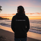 Man wearing a black Deepwater Crew Co. performance UPF hoodie standing on a beach at sunset.