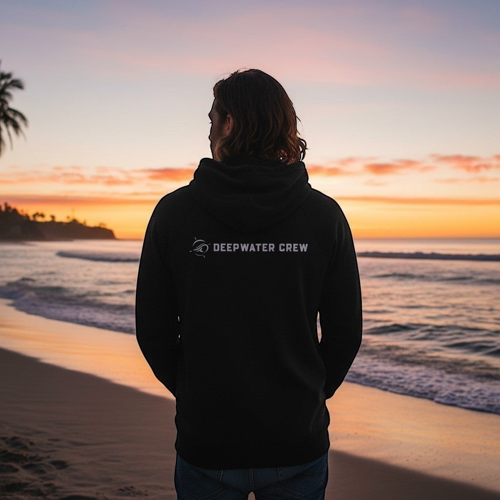 Man wearing a black Deepwater Crew Co. performance UPF hoodie standing on a beach at sunset.