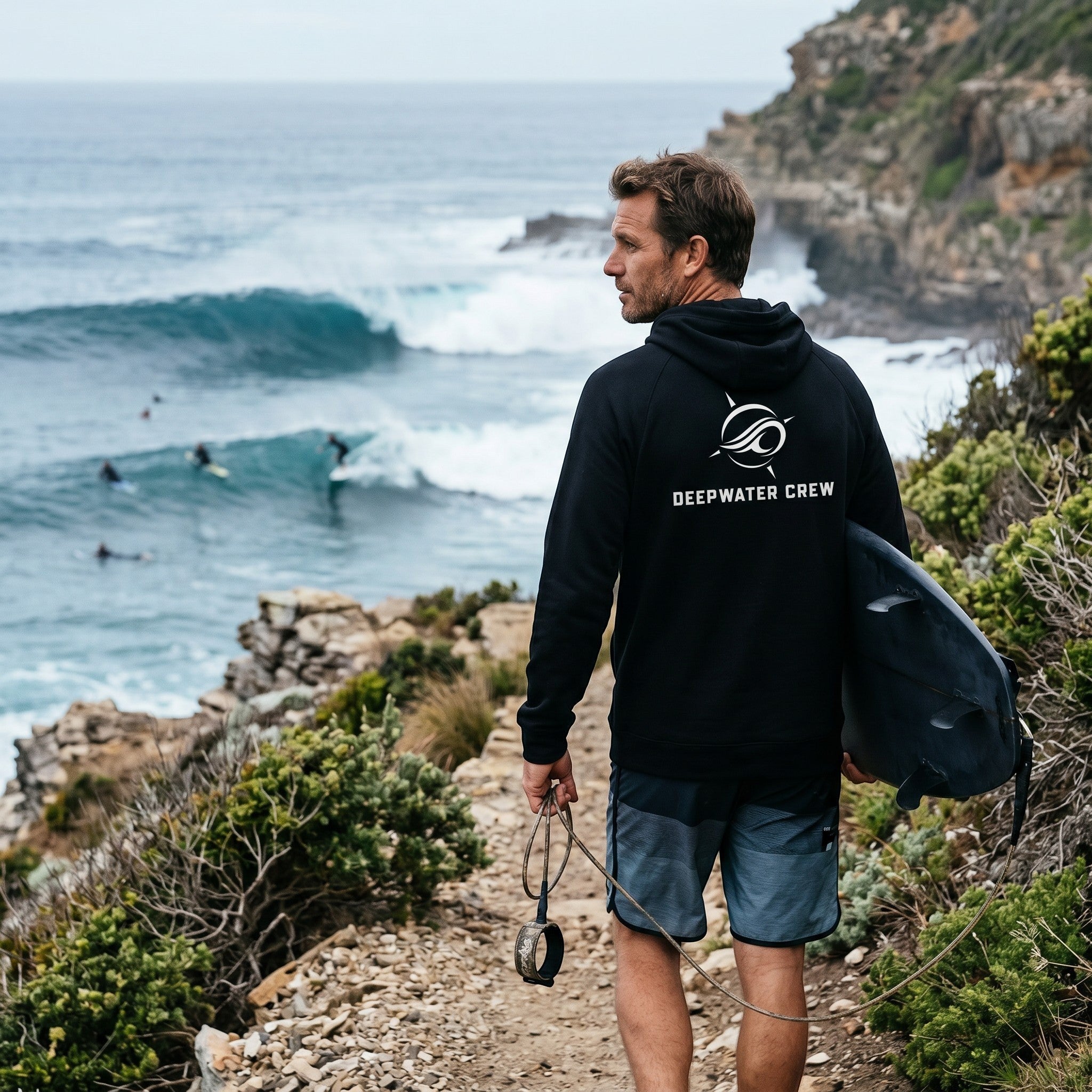 Man wearing a black Deepwater Crew Co. performance UV hoodie with logo, standing on a cliff overlooking the ocean.