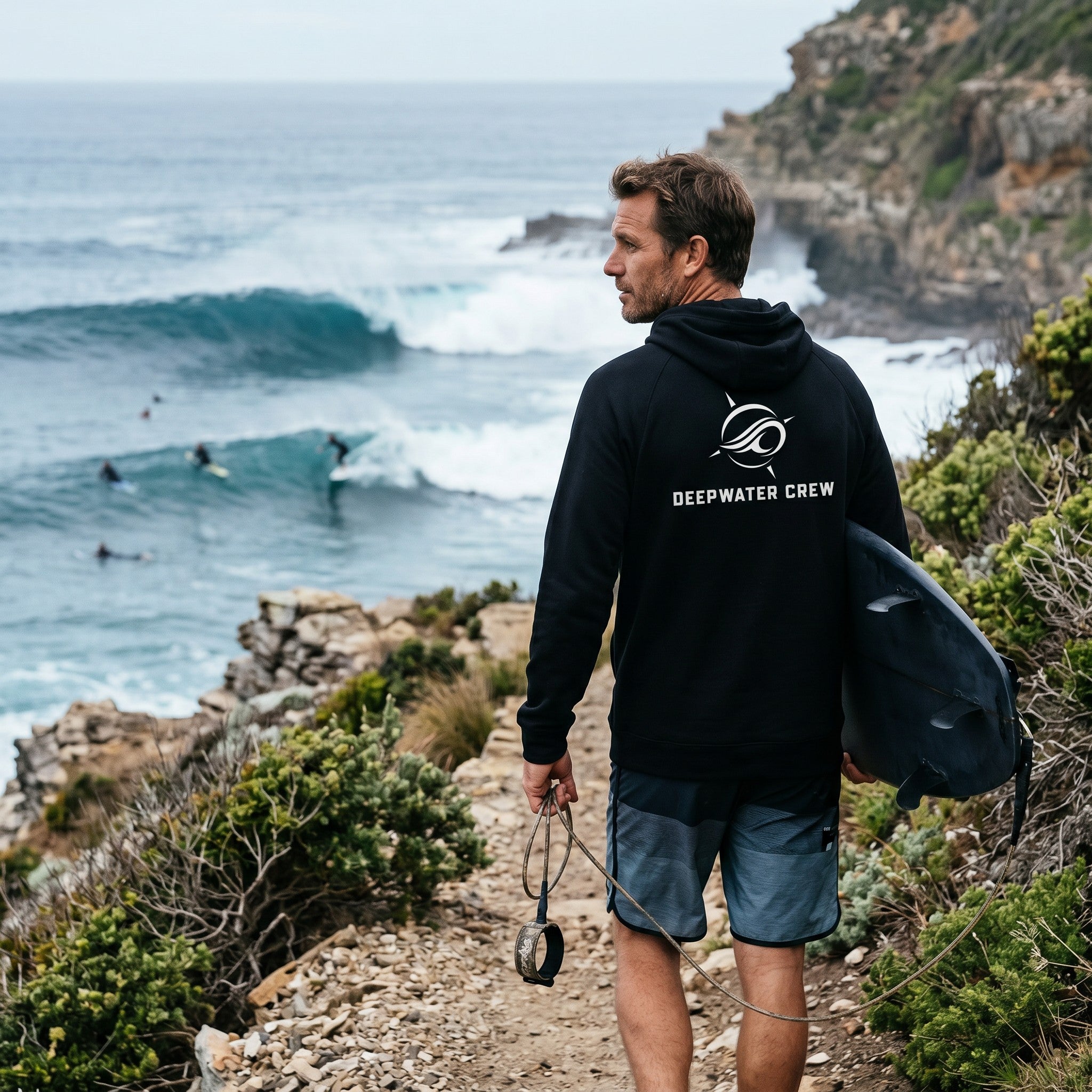 Deepwater Crew Co. man wearing a black performance UV hoodie with logo, holding a surfboard on a coastal cliff path.