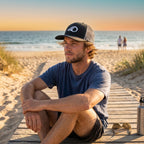Deepwater Crew Co. black trucker hat with white wave logo worn by a man sitting on a beach boardwalk at sunset