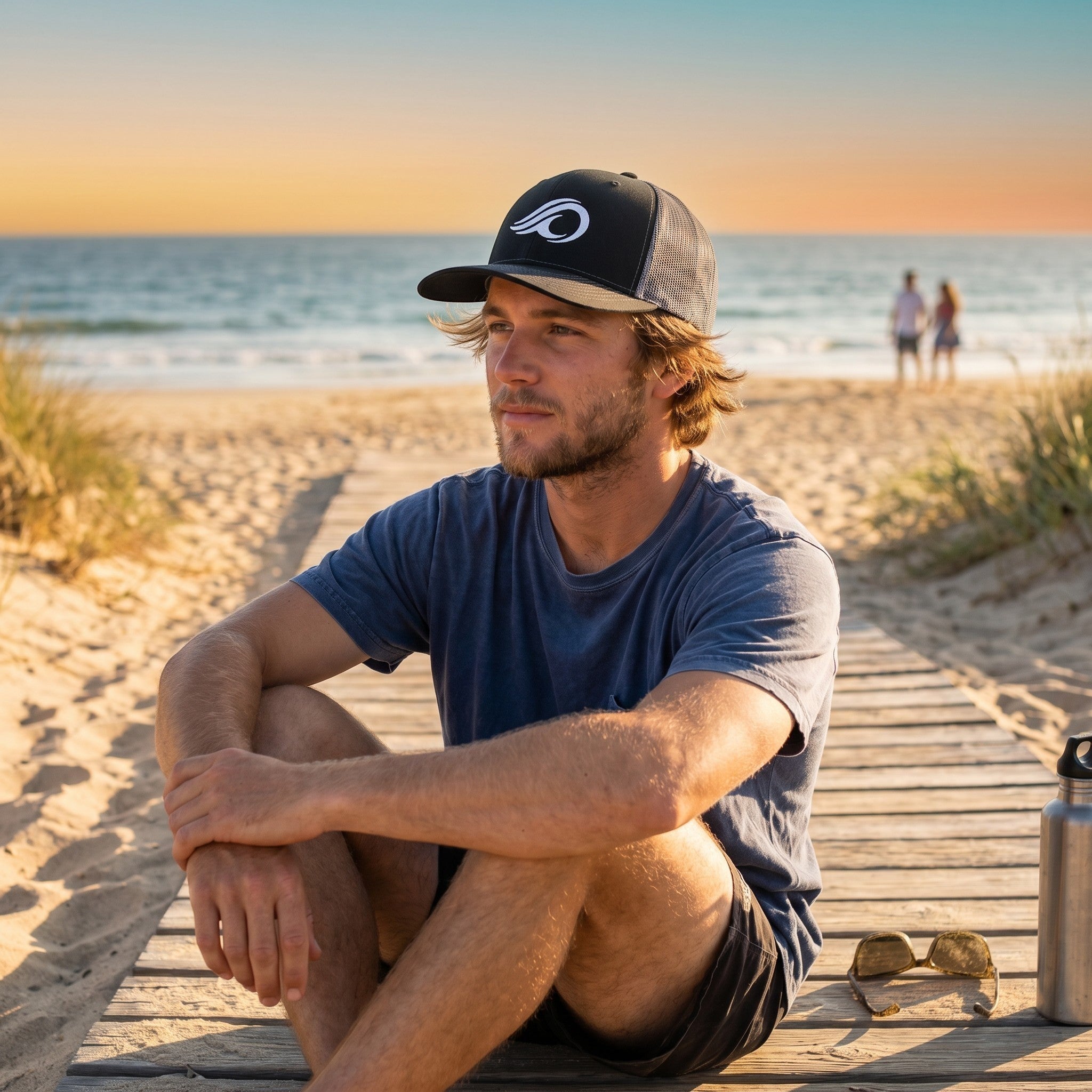 Deepwater Crew Co. black trucker hat with white wave logo worn by a man sitting on a beach boardwalk at sunset