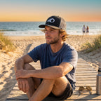Deepwater Crew Co. black trucker hat with white wave logo worn by a man sitting on a beach boardwalk at sunset