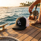 Deepwater Crew Co. black and white trucker hat with embroidered logo on a wooden boat deck at sunset.