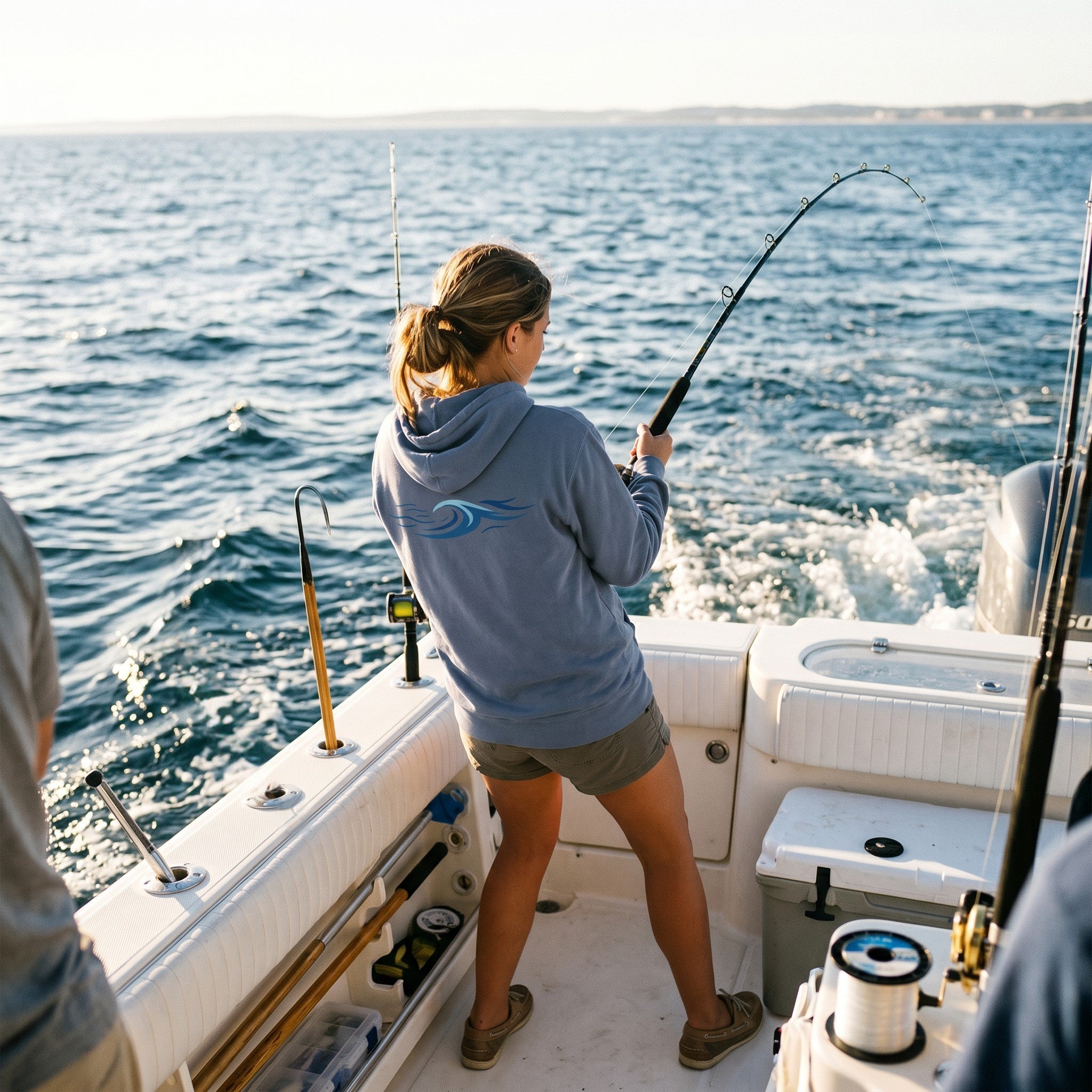Woman wearing a Deepwater Crew Co. blue hooded long sleeve tee with a wave logo while fishing on a boat at sea.