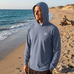 Man wearing a blue Deepwater Crew Co. long sleeve hooded tee on a sandy beach at sunset.