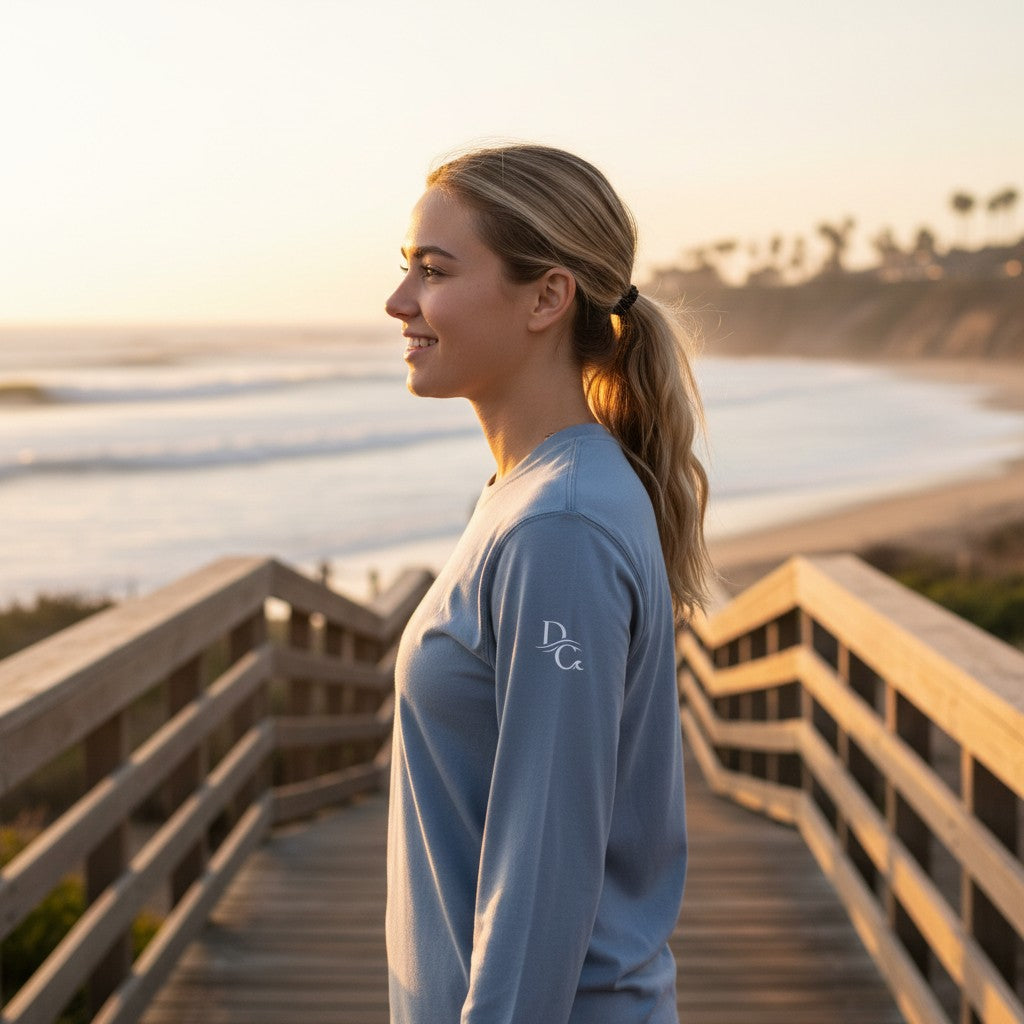Woman wearing a light blue Deepwater Crew Co. hooded long sleeve tee on a wooden boardwalk at sunset.