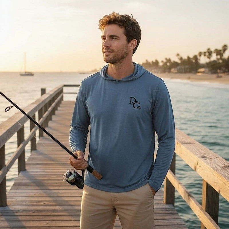 Man wearing a blue Deepwater Crew Co. long sleeve hoodie tee with a fishing rod on a wooden pier at sunset