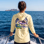 Woman wearing a pale yellow Deepwater Crew Co. Bluewater Strike UPF 50+ performance hoodie, back view with colorful pelagic game fish collage and Deepwater Crew lettering while boating on open water.
