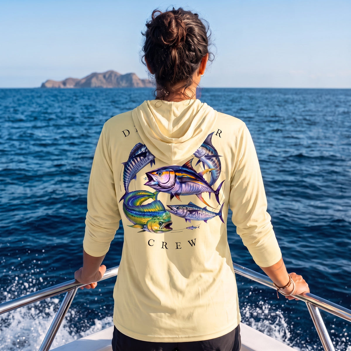 Woman wearing a pale yellow Deepwater Crew Co. Bluewater Strike UPF 50+ performance hoodie, back view with colorful pelagic game fish collage and Deepwater Crew lettering while boating on open water.