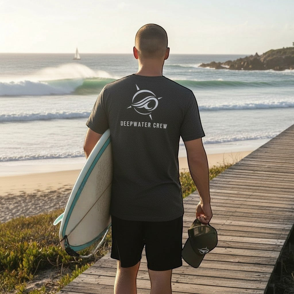 Man wearing a Deepwater Crew Co. charcoal grey cotton t-shirt with a white logo on the back, holding a surfboard.