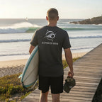 Man wearing a Deepwater Crew Co. charcoal grey cotton t-shirt with a white logo on the back, holding a surfboard.