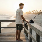 Deepwater Crew Co. cream cotton t-shirt with back graphic, worn by a man fishing on a wooden pier at sunset.