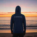 Deepwater Crew Co. dark blue performance UPF hoodie with logo, worn by a person facing the ocean at sunset.