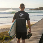 Deepwater Crew Co. dark grey crew neck t-shirt with logo on back, worn by a man holding a surfboard.