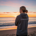 Deepwater Crew Co. dark grey performance UPF hoodie worn by a woman looking at a sunset over the ocean.