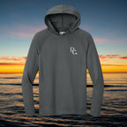 Deepwater Crew Co. dark grey performance UV hoodie with raglan sleeves and white logo against a sunset ocean backdrop.