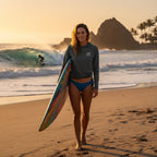 Deepwater Crew Co. dark grey performance UPF hoodie worn by a woman holding a surfboard on a sunny beach at sunset.