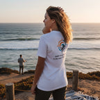 Woman standing by the ocean in a white Deepwater Crew Co. Deep 2 Deep T-shirt, back view showing a colorful compass sun and wave graphic above Deep Calls to Deep Psalm 42 text.