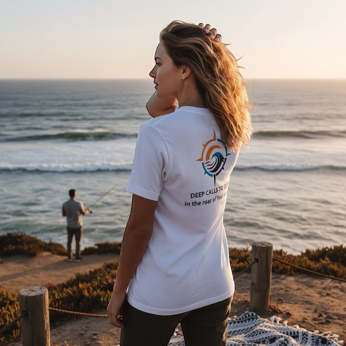 Woman standing by the ocean in a white Deepwater Crew Co. Deep 2 Deep T-shirt, back view showing a colorful compass sun and wave graphic above Deep Calls to Deep Psalm 42 text.