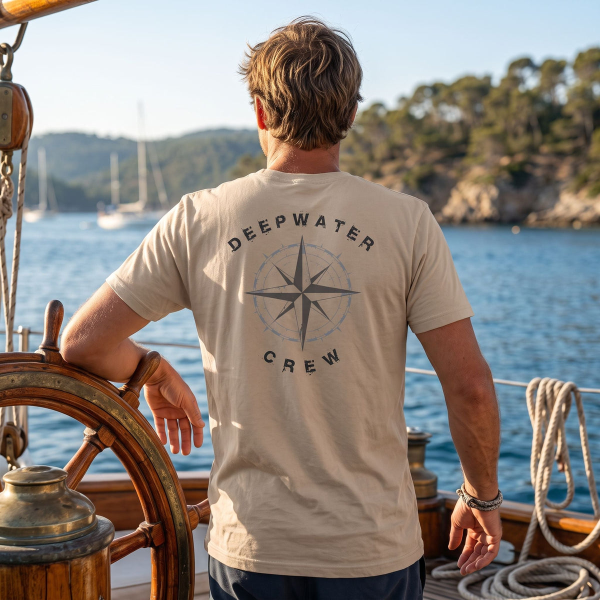 Man wearing a tan Deepwater Crew Co. Deepwater Nomad crewneck T-shirt, back view with distressed compass rose graphic while standing at the helm of a sailboat.
