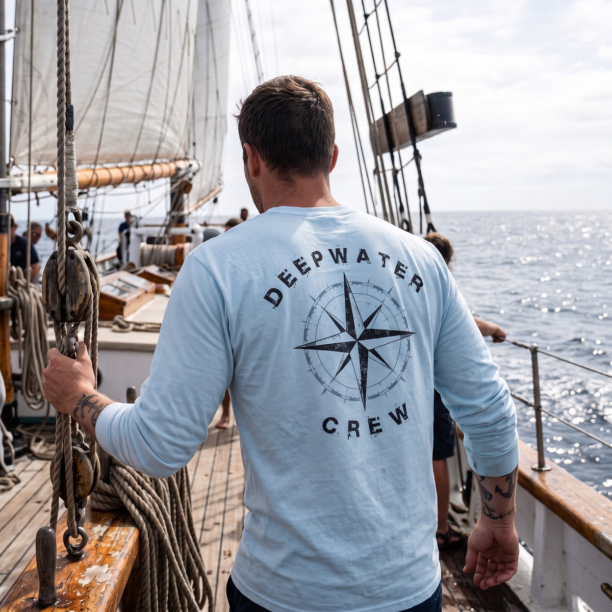 Man wearing a light blue Deepwater Crew Co. Deepwater Nomad UPF 44+ long-sleeve performance shirt, back view with distressed compass rose graphic while sailing on open water.