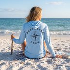 Woman wearing a Blue Mist Deepwater Crew Co. Deepwater Nomad UPF 50+ performance hoodie, back view with distressed compass rose graphic while relaxing on the beach.