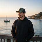 Deepwater Crew Co. green and black trucker hat with embroidered logo, worn by a man on a pier at sunset.