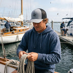 Deepwater Crew Co. grey and black trucker cap with wave logo worn by a man on a boat at a marina.