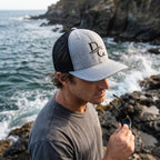 Deepwater Crew Co. grey and black mesh trucker cap with embroidered logo worn by a man on a rocky coastline