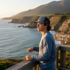 Deepwater Crew Co. grey and black trucker hat with embroidered logo, worn by a man fishing by the ocean.