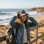 Woman wearing a Deepwater Crew Co. grey and black trucker hat with a wave logo at a coastal beach.