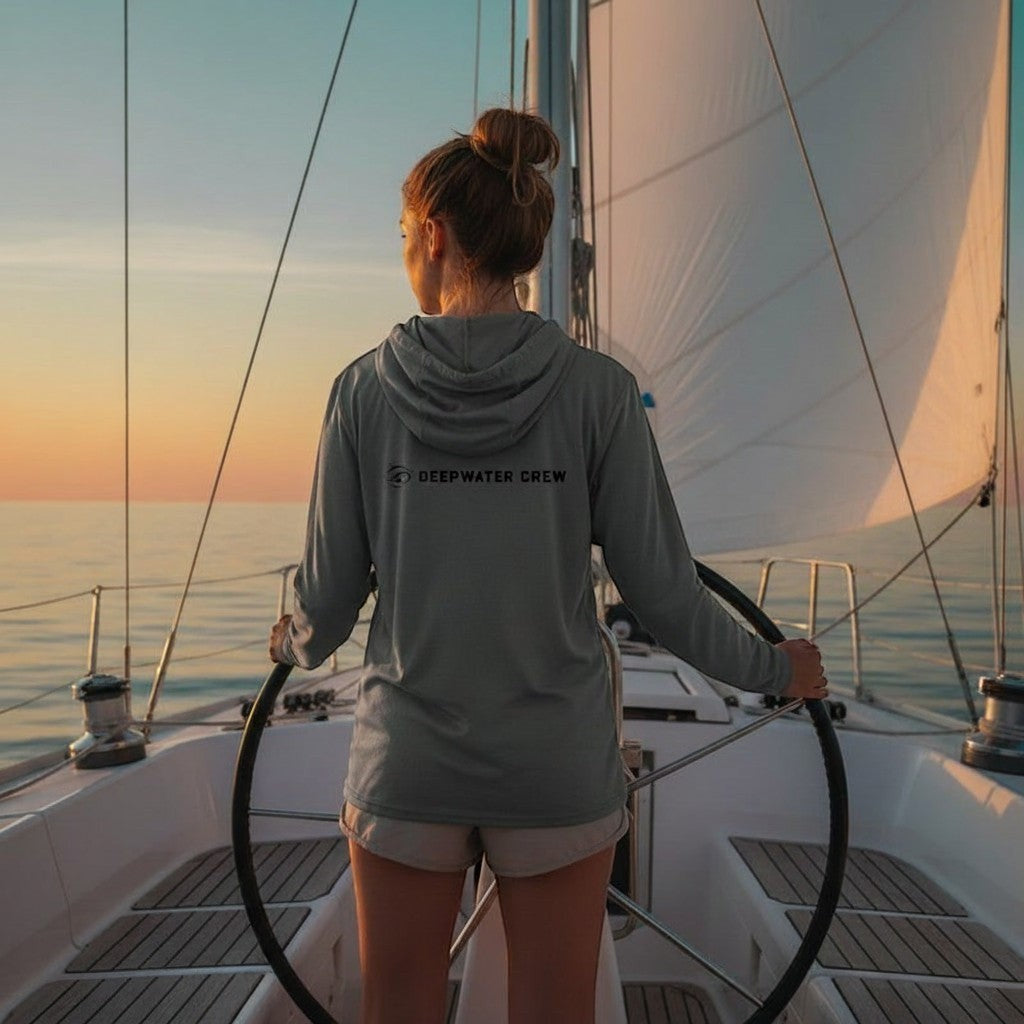 Deepwater Crew Co. grey tri-blend long-sleeve hooded tee with chest logo, worn by a woman on a sailboat.