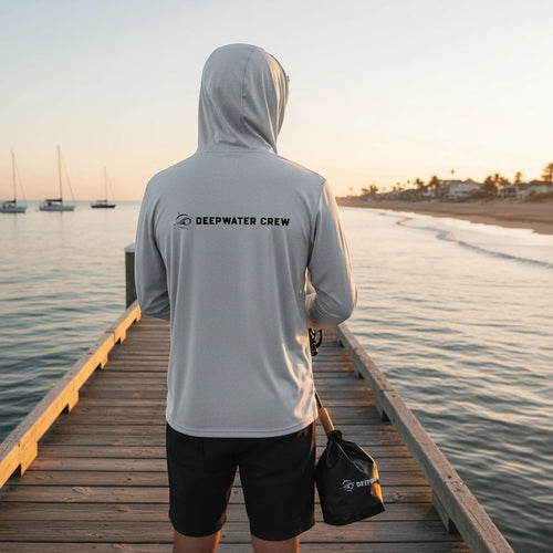 Deepwater Crew Co. light grey hooded long-sleeve tee with chest logo, worn by a man fishing on a pier at sunset.
