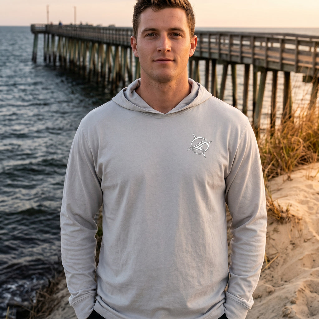 A man wearing a Deepwater Crew Co. light grey long-sleeve hooded tee with a small logo on the chest by the ocean.