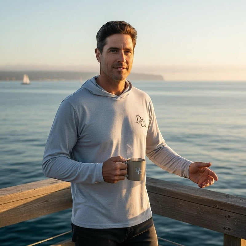 Deepwater Crew Co. man wearing a light grey long sleeve tri-blend hoodie with a chest logo on a coastal deck at sunset.
