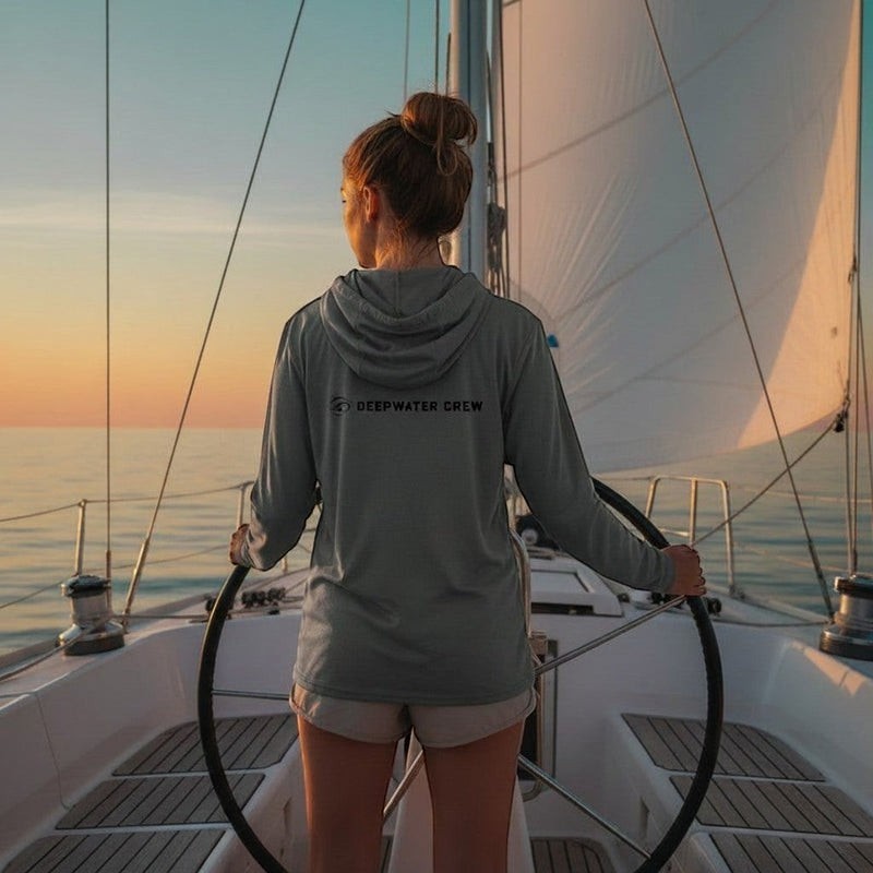 Deepwater Crew Co. grey long sleeve hoodie with logo on back, worn by a woman steering a sailboat at sunset.