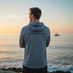Deepwater Crew Co. man wearing a grey tri-blend long sleeve hoodie with a logo, standing by the ocean at sunset.