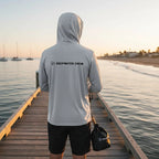 A man wearing a Deepwater Crew Co. grey long sleeve hoodie with a logo on the back while standing on a wooden pier.