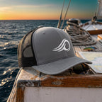 Deepwater Crew Co. grey and black mesh trucker hat with white wave logo embroidery on a boat deck at sunset.
