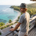 Deepwater Crew Co. grey trucker cap with flat bill and embroidered logo worn by a man overlooking a tropical beach